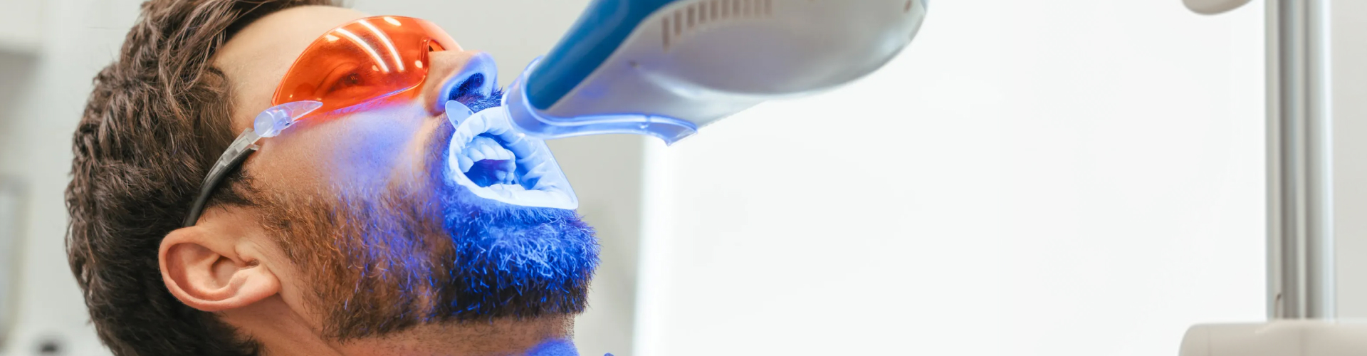 Gloved hand shining blue light on man's teeth wearing orange glasses.